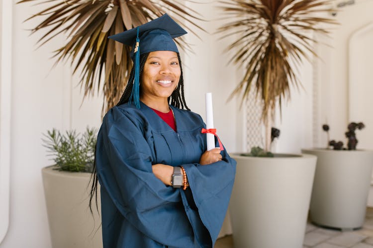 Woman In Academic Gown Holding A Rolled Certificate While Smiling At The Camera