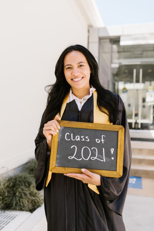 Free Proud graduate in gown holding Class of 2021 sign outdoors, smiling. Stock Photo