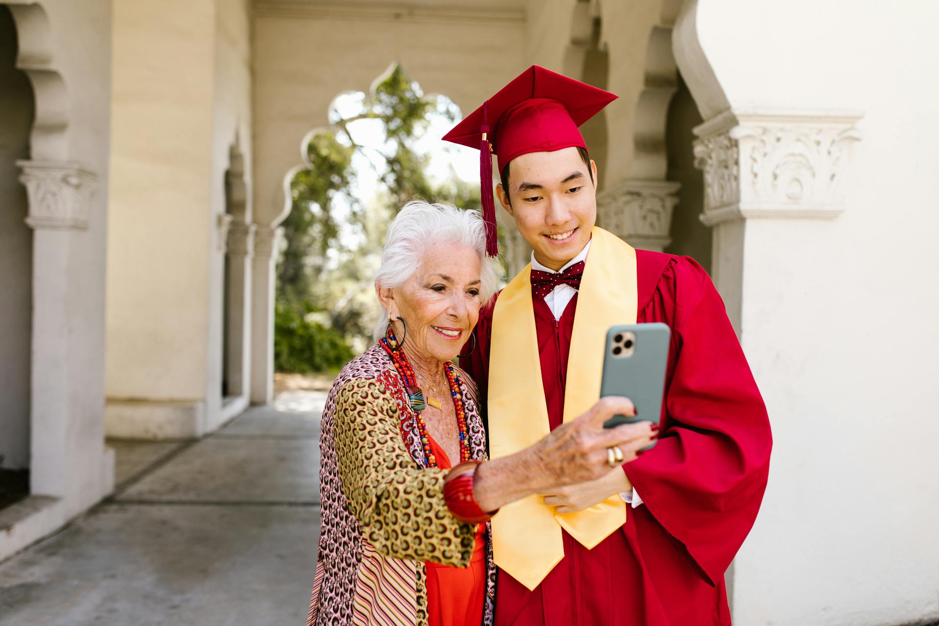 Young graduate in cap and gown takes a joyful selfie with proud grandmother outside university building.