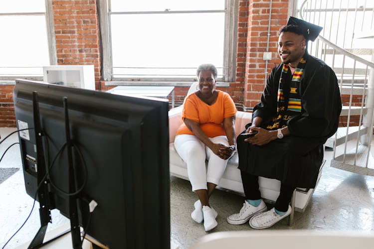 Man In A Graduate Gown And Cap Playing Video Games With His Mother