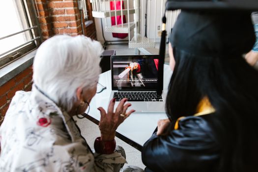 A senior and a graduate watch a virtual graduation ceremony on a laptop at home.