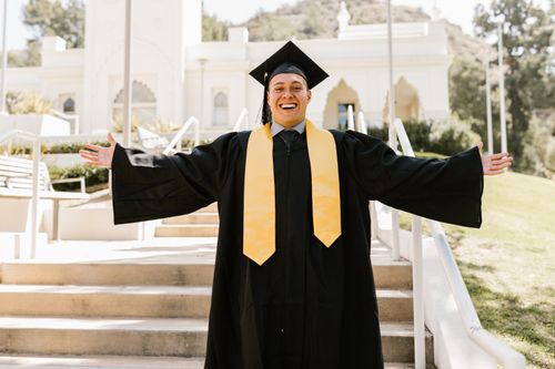 Free A joyful graduate in cap and gown celebrates outdoors with arms open wide, expressing achievement and happiness. Stock Photo
