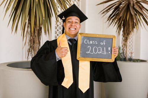 Free Cheerful young graduate in cap and gown holding Class of 2021 sign with thumbs up. Stock Photo