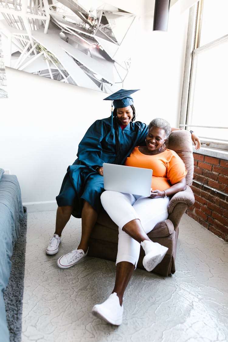 Mother And Daughter Sitting On An Armchair Watching Virtual Graduation Ceremony On Laptop