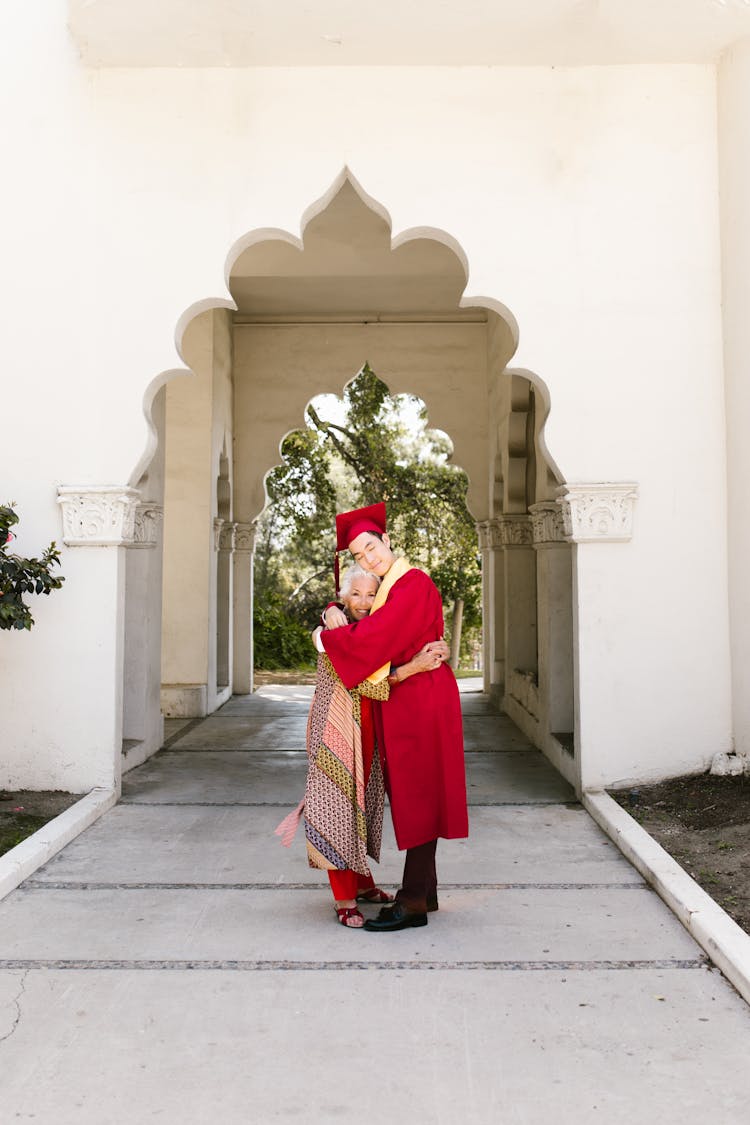 Student In Red Graduation Gown Hugging The Elderly Woman 