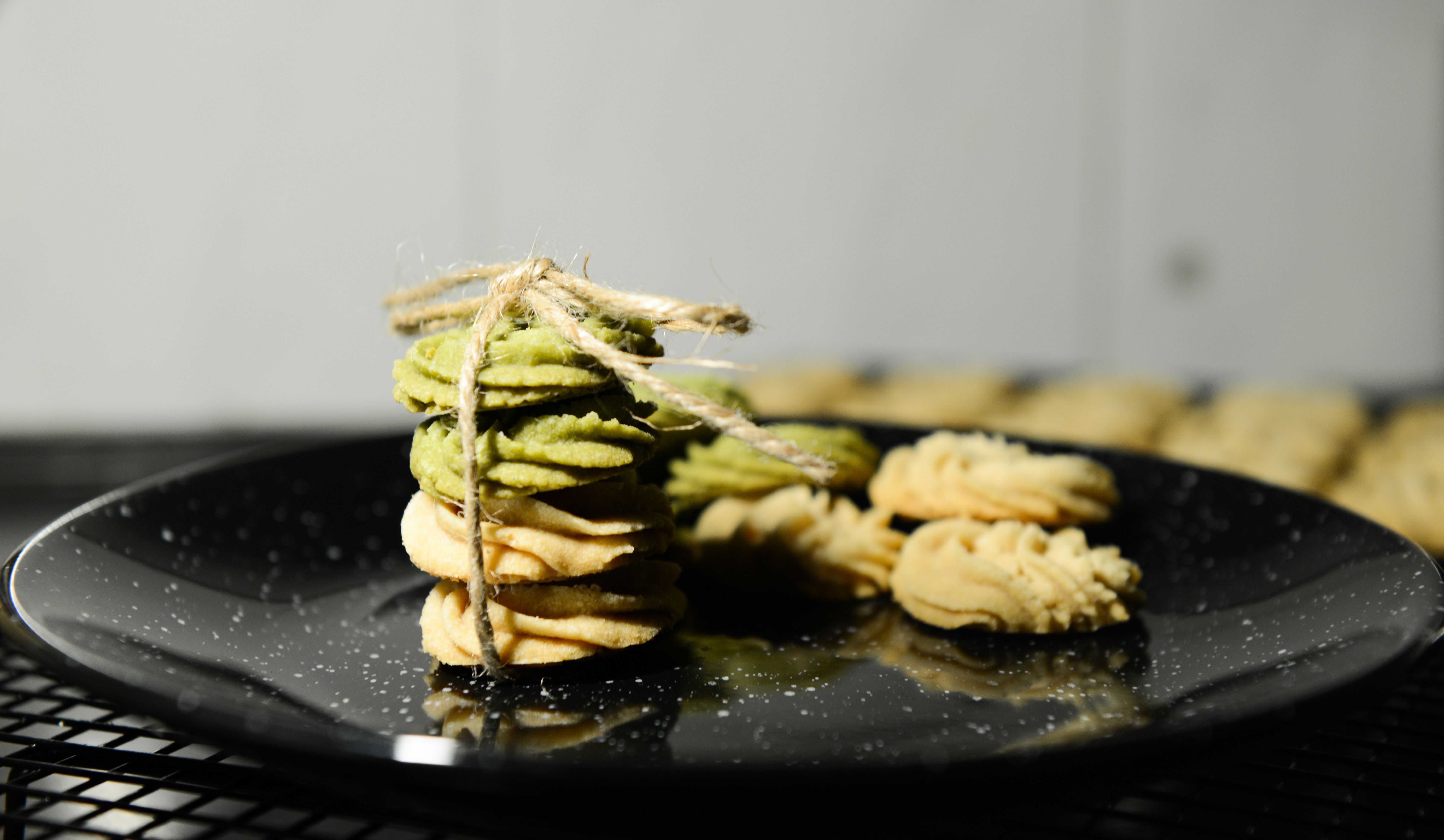A plate of pastel-colored star and wreath-shaped spritz cookies dusted with colored sugar, stacked next to a metal cookie press.