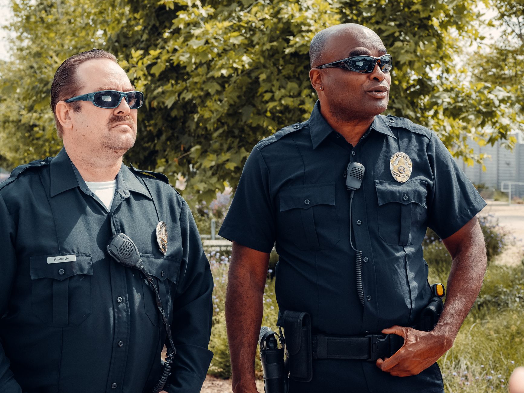 Two police officers in uniform, wearing sunglasses, standing outdoors with a serious demeanor.
