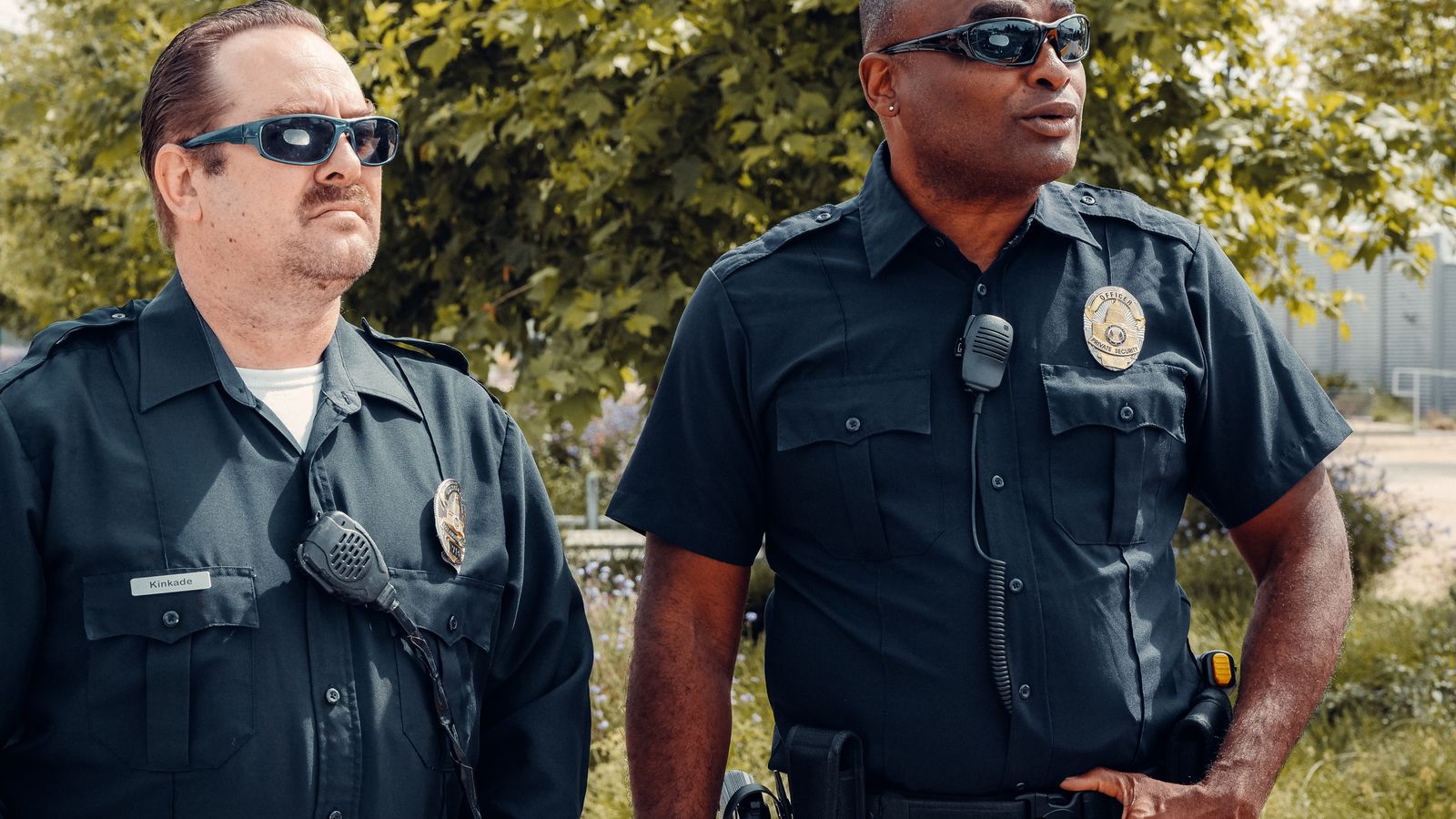 Two police officers in uniform, wearing sunglasses, standing outdoors with a serious demeanor.