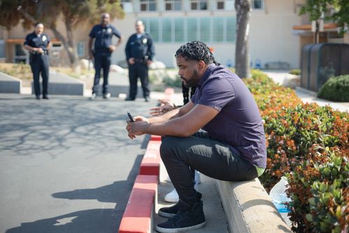 Free A man sits on a curb using a smartphone with officers in the background. Stock Photo