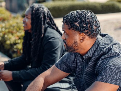 Two men with braided hairstyles sitting outdoors, captured in a close-up highlighting style and focus.
