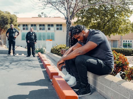 Police officers observing two adults sitting on a curb, possibly detained, in a sunny outdoor setting.