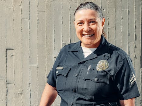 A cheerful policewoman smiling confidently in her uniform against a concrete wall.