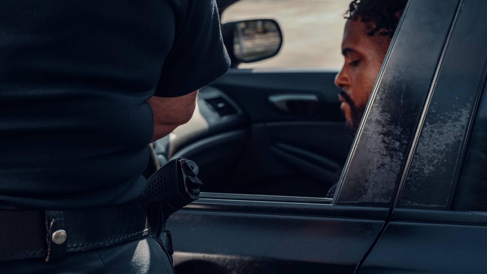 A police officer issuing a ticket to a man seated in a car, captured outdoors in daylight.