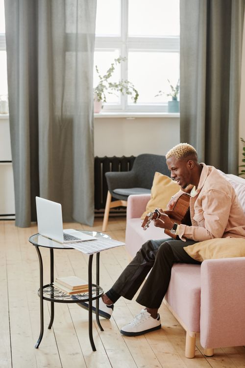 Free Young man playing acoustic guitar while sitting on a sofa during a video call at home. Stock Photo