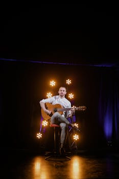 Musician playing acoustic guitar on stage, surrounded by warm stage lights.