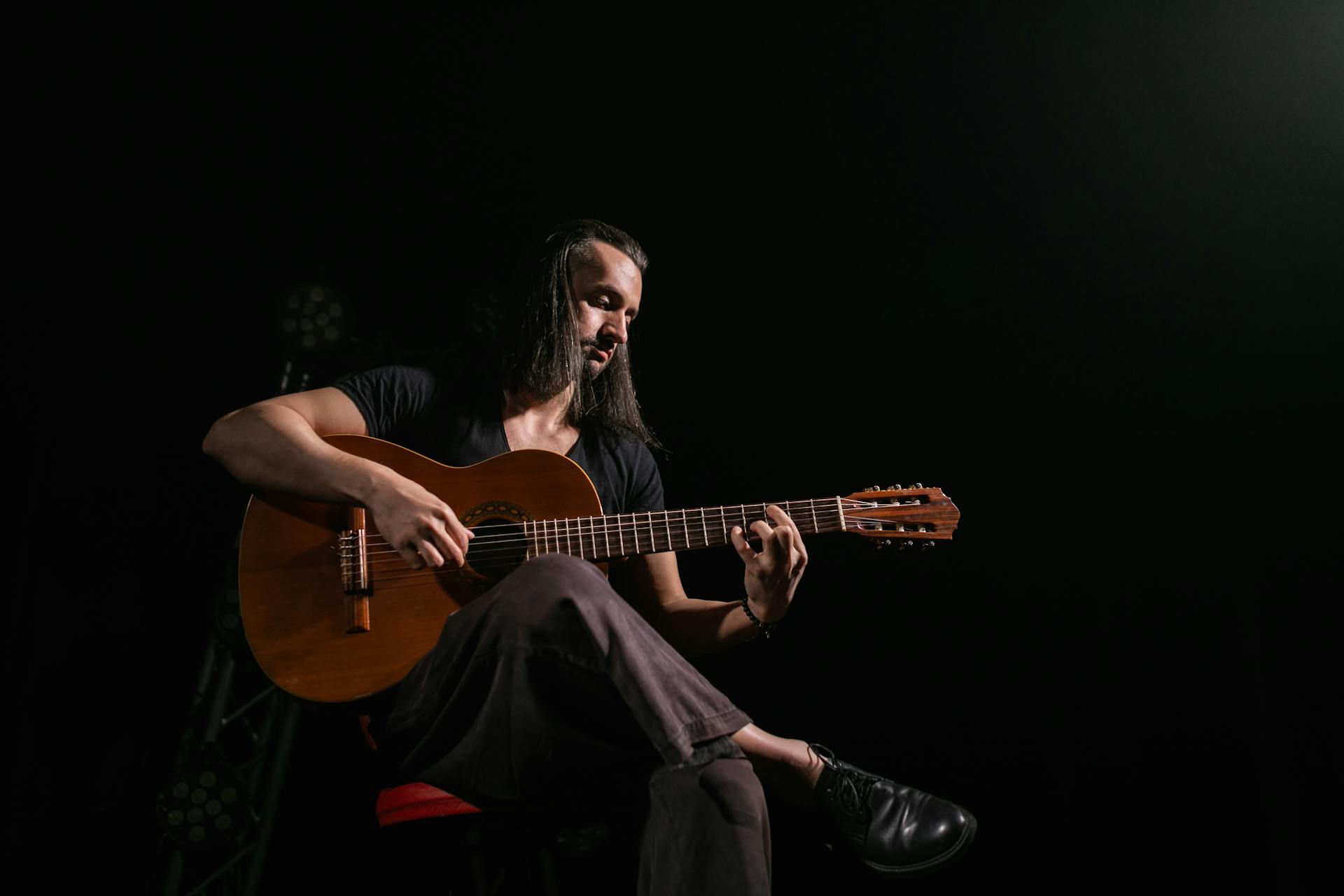 A musician with long hair sitting and playing a guitar in a dark room.