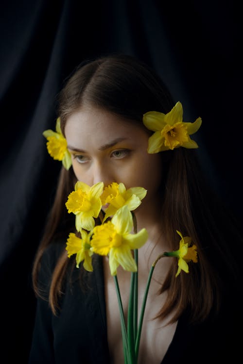 Young sensitive woman with brown hair covering mouth with blossoming yellow Narcissus flowers while looking down