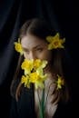Young sensitive woman with brown hair covering mouth with blossoming yellow Narcissus flowers while looking down