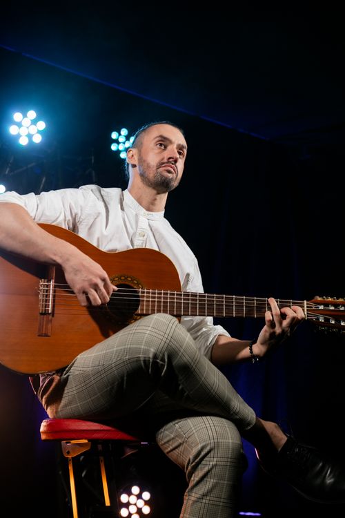 Free A bearded man in white long sleeves plays an acoustic guitar on stage under dramatic lighting. Stock Photo