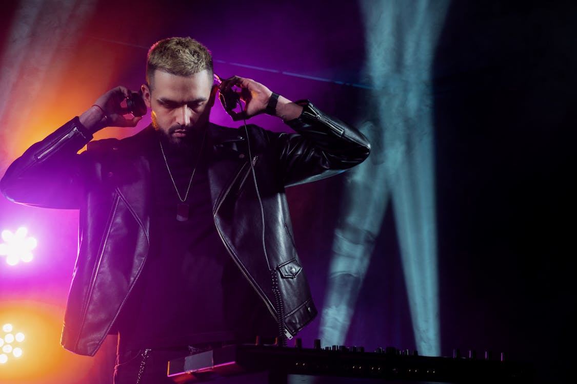 Free Self assured young bearded male DJ in trendy leather jacket adjusting headphones while performing music on stage in dark illuminated nightclub Stock Photo