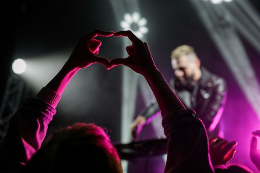 A lively concert scene featuring a heart-shaped hand gesture against a vivid purple backdrop.