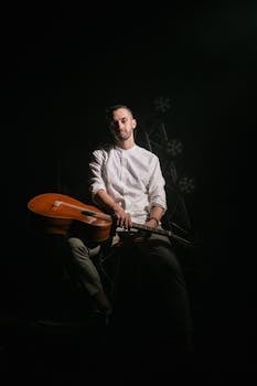 A male musician holding an acoustic guitar, seated on stage with dramatic lighting.