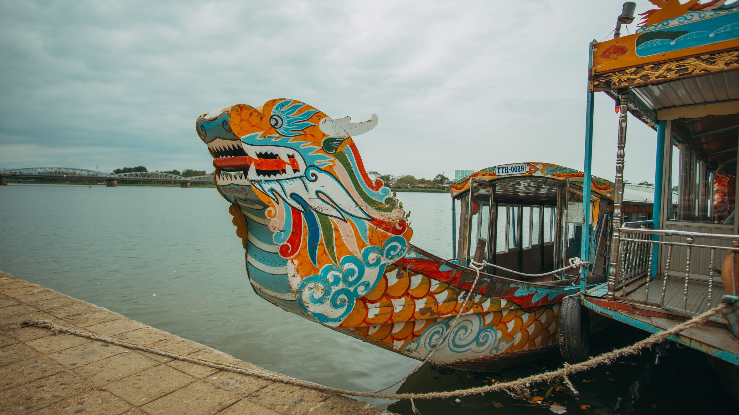 A vibrant dragon boat docked on a riverfront under cloudy skies, showcasing intricate art.