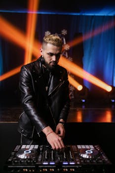 DJ in a black leather jacket operating a mixer in a nightclub with vibrant spotlights.