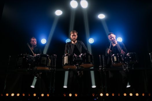 Three male musicians perform on stage with drums under dramatic lighting.
