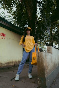 Woman in casual yellow attire and bucket hat posing outdoors with a tote bag.