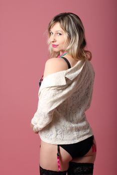 Back view of young sensual blond haired female model in black stockings wearing black lingerie and white blazer standing against pink background and looking at camera over shoulder