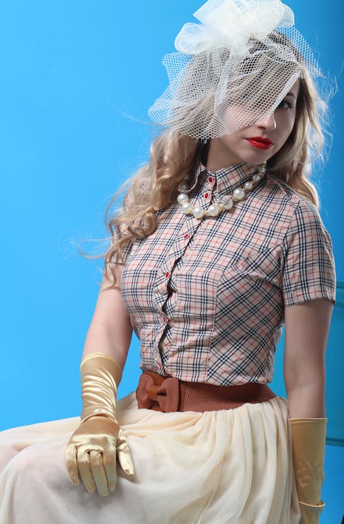 Young blond haired female model with bright makeup wearing elegant skirt and checkered shirt with evening gloves and white hat with covered face veil standing against blue background