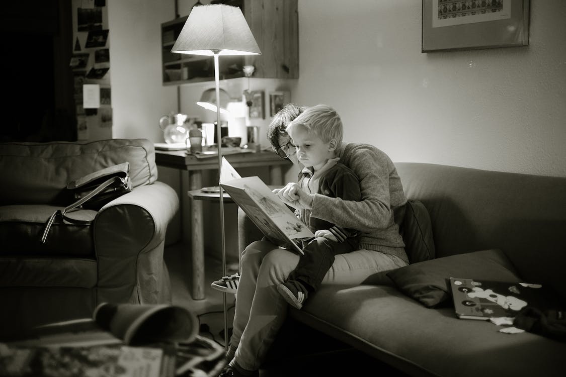 Grayscale Photography of Child on Lap of Woman While Reading Book