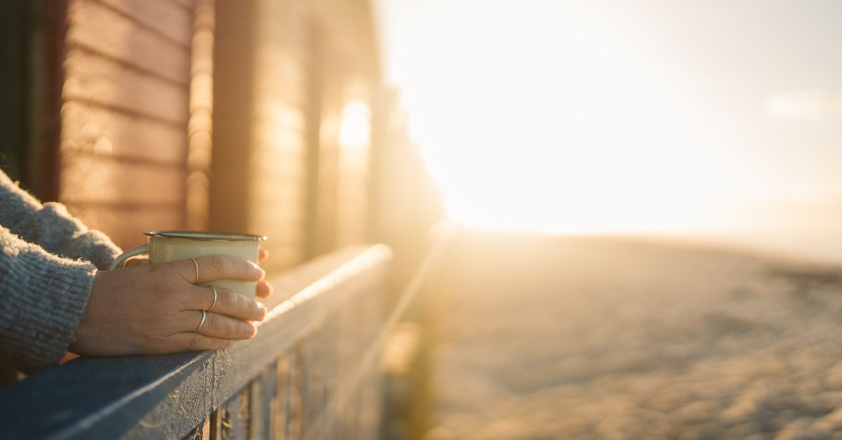 A person holding a simple ceramic mug of coffee while looking out a window at sunrise, with no phone in sight