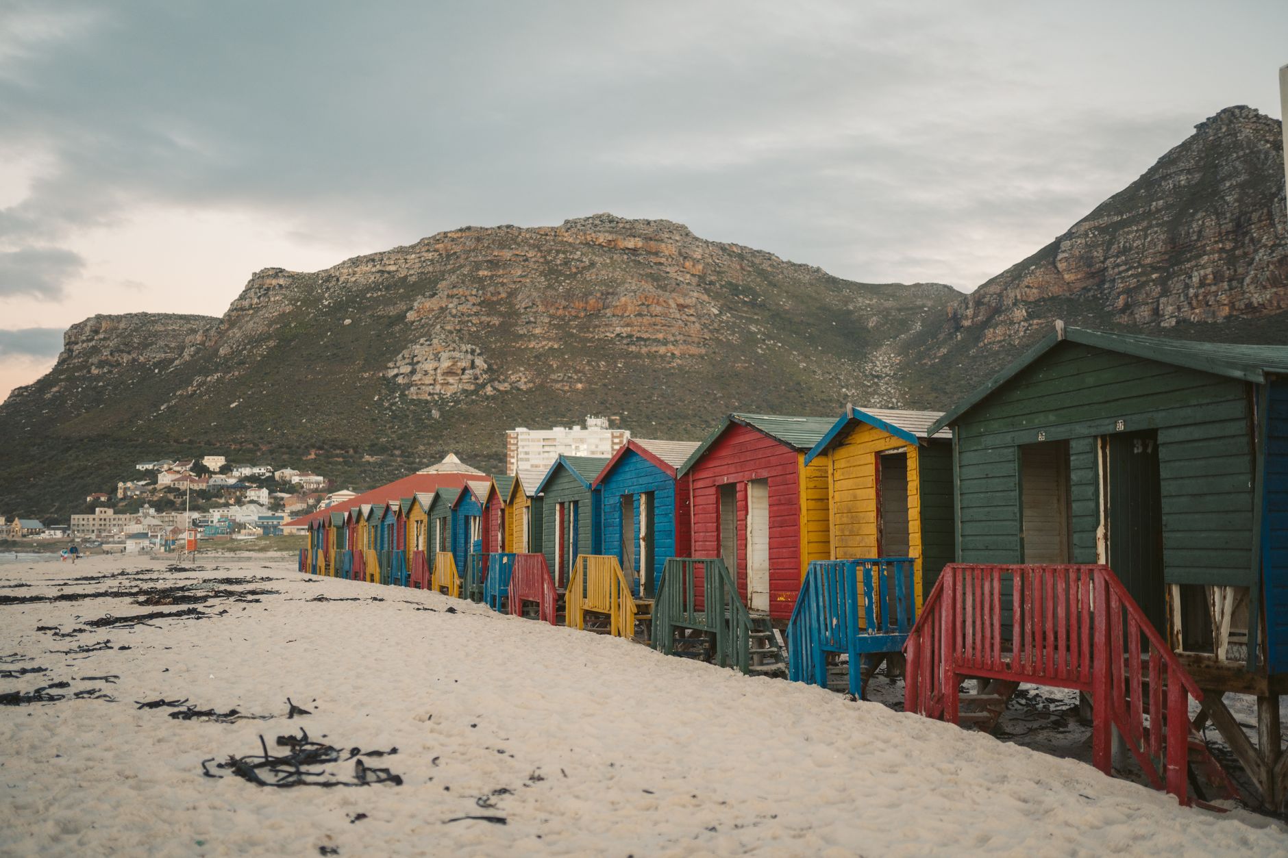 Vibrant wooden beach huts on sandy Muizenberg Beach with mountains in the background.