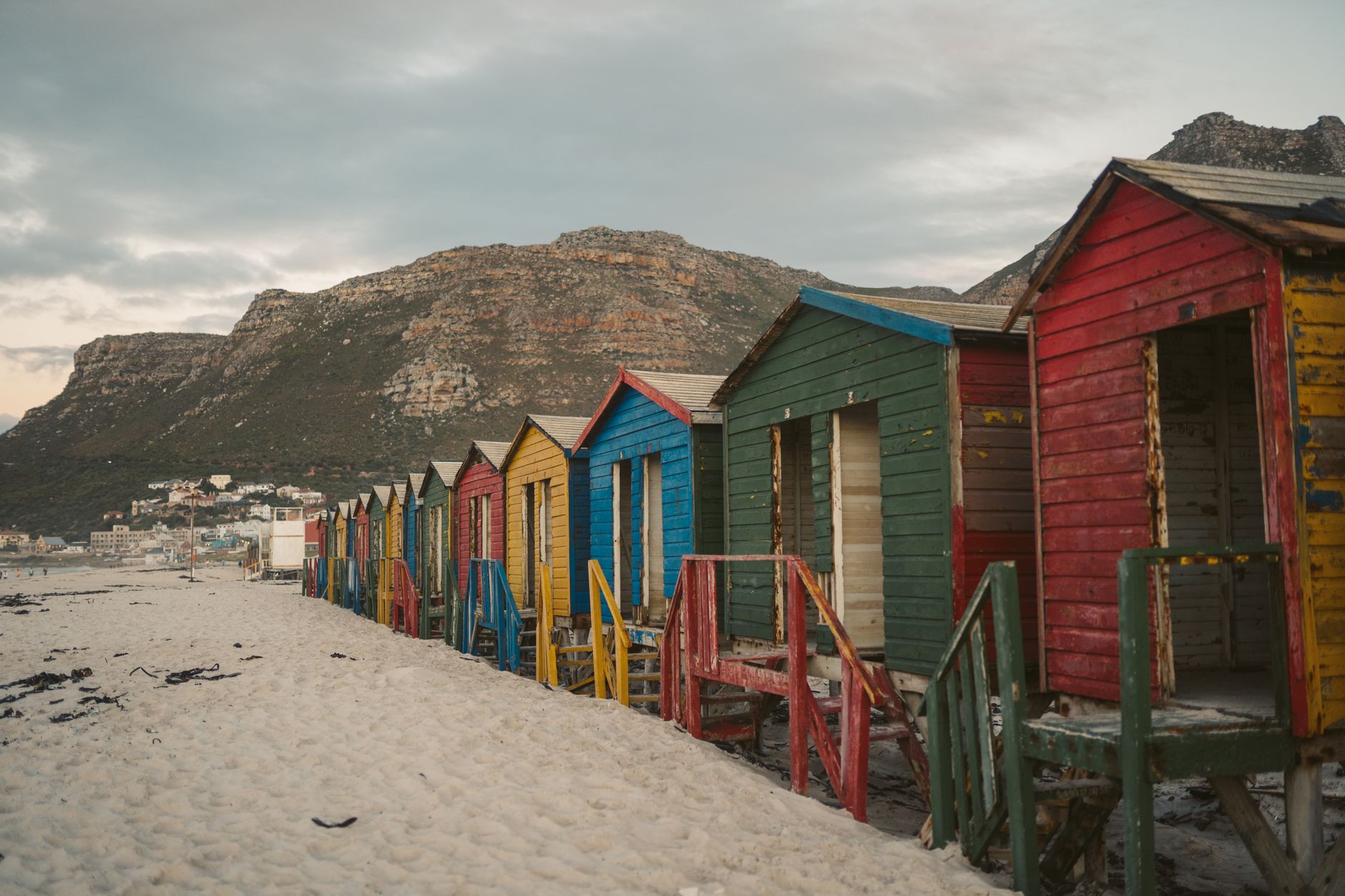 Colorful wooden beach huts line a sandy shore, backed by mountains and a cloudy sky.