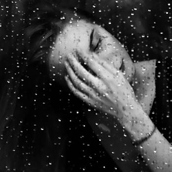 Black and white portrait of a woman behind a rain-soaked window, evoking solitude.