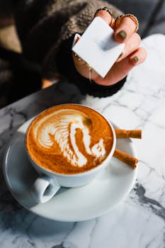 Close-up of a latte with intricate art design on a marble table with a hand holding sugar.