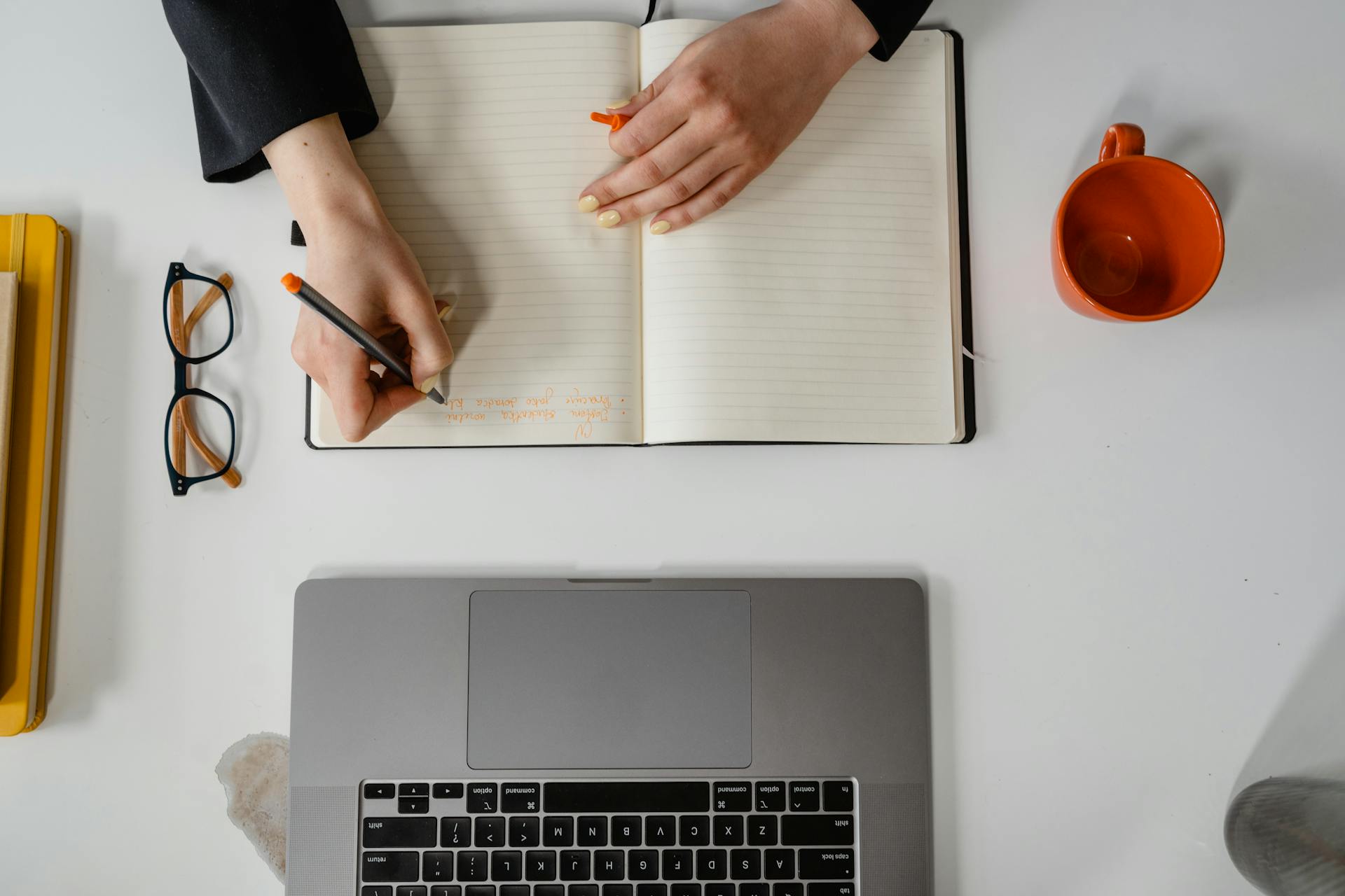 A businesswoman writes notes in a notebook with a laptop, glasses, and mug in view.