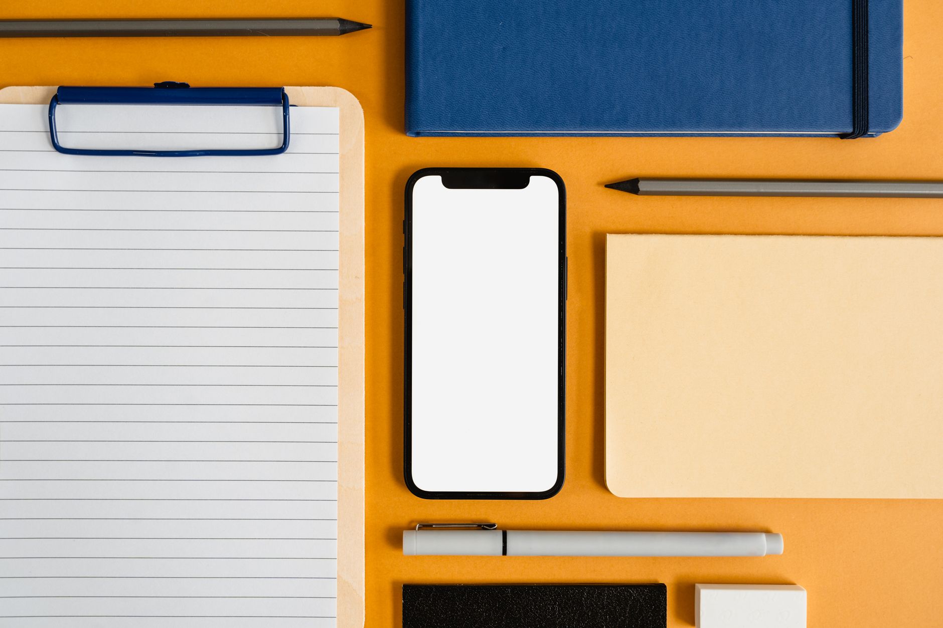 A top view of office essentials including a smartphone, clipboard, and stationery on a yellow surface.
