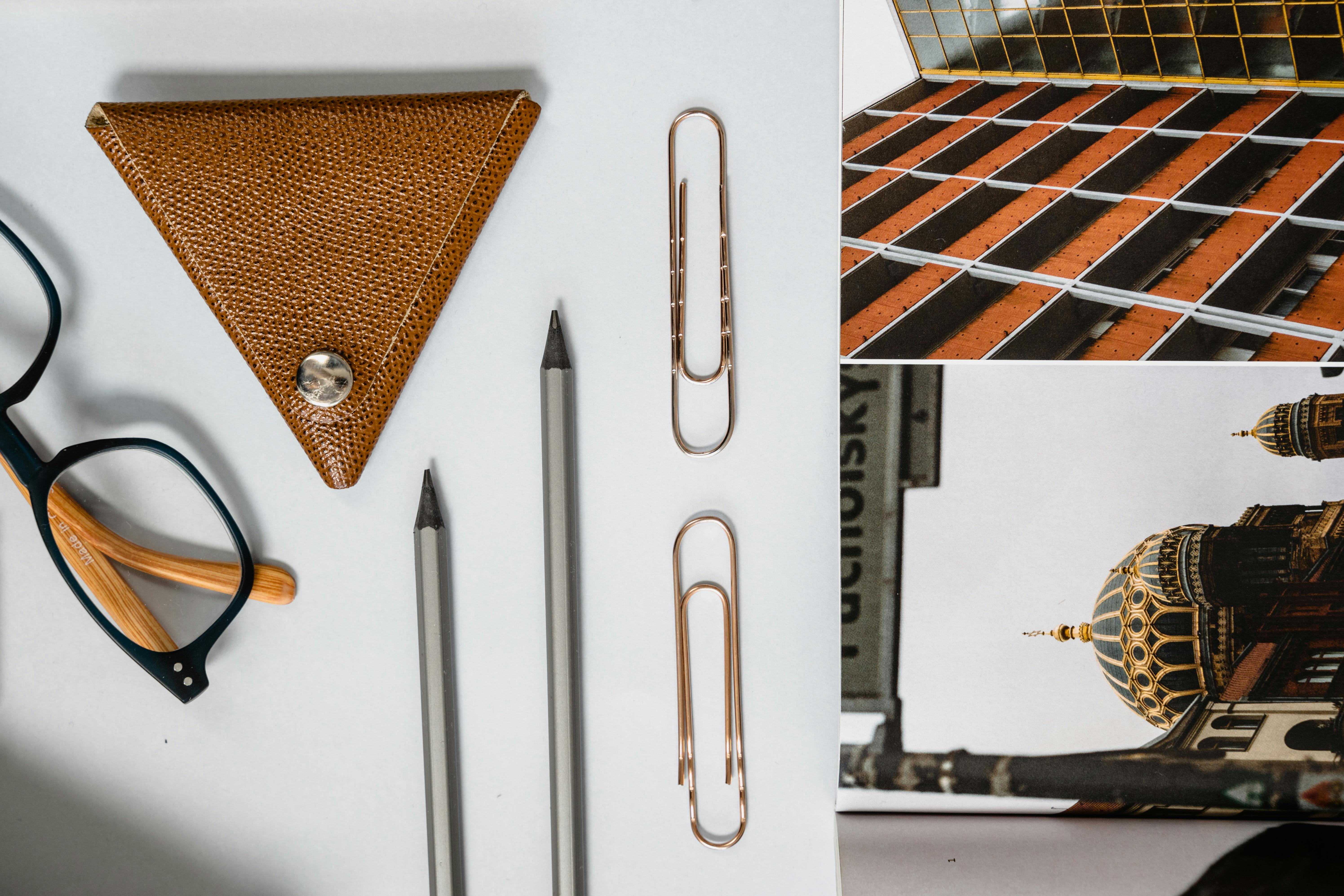 Flat lay of eyeglasses, pencils, paper clips, and leather keyholder on a white background with architectural postcard.