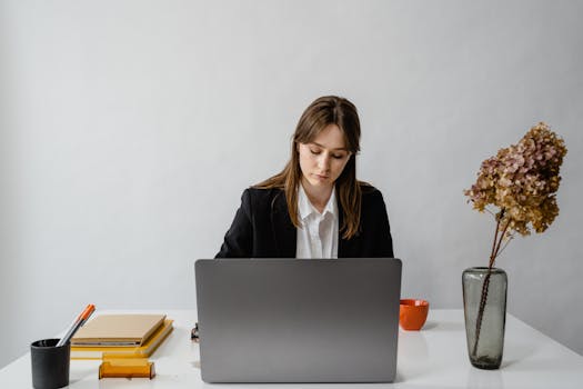 Professional woman working at a white desk with a laptop, stationery, and dried flowers in an office setting.
