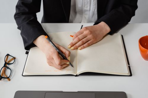 Free A person writing in a notebook at a desk with eyeglasses and a laptop nearby. Stock Photo