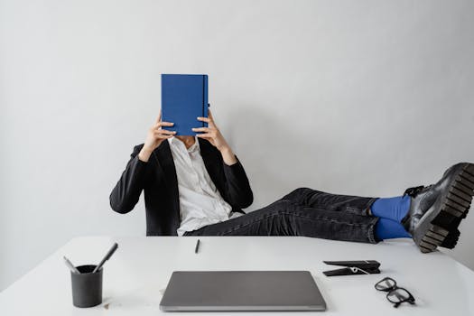 Man in casual office setting reading a blue notebook with feet up on desk.