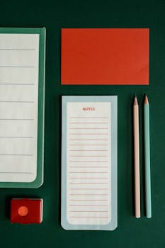 Artistic arrangement of office stationery with a notepad, pencils, and red accents on a green backdrop.