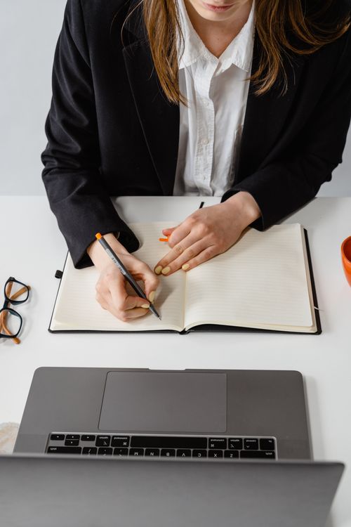 Free Overhead view of a professional woman in a black blazer writing in a notebook at her desk. Stock Photo