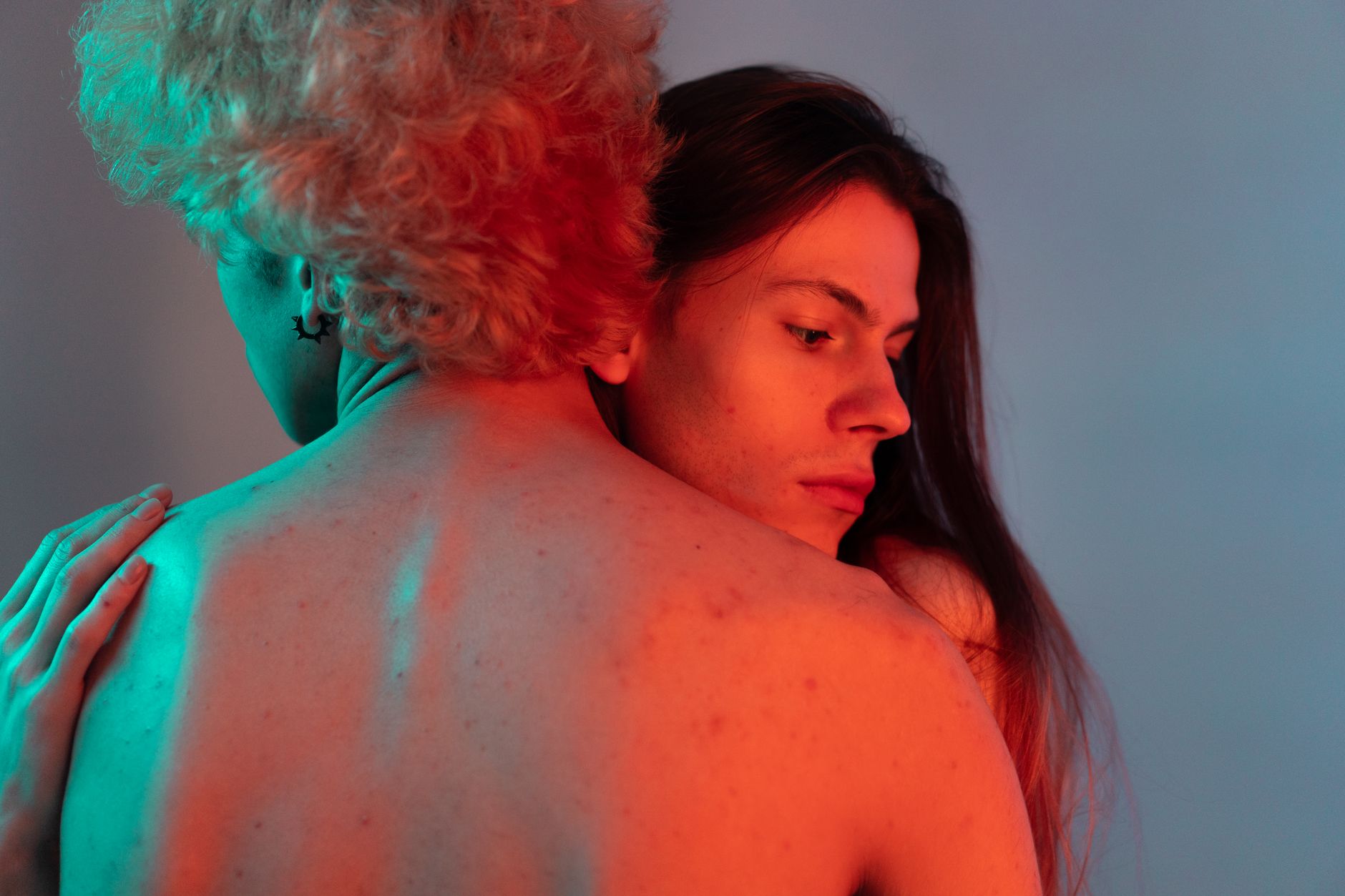 A romantic studio portrait of a topless LGBT couple embracing, bathed in colorful lighting.