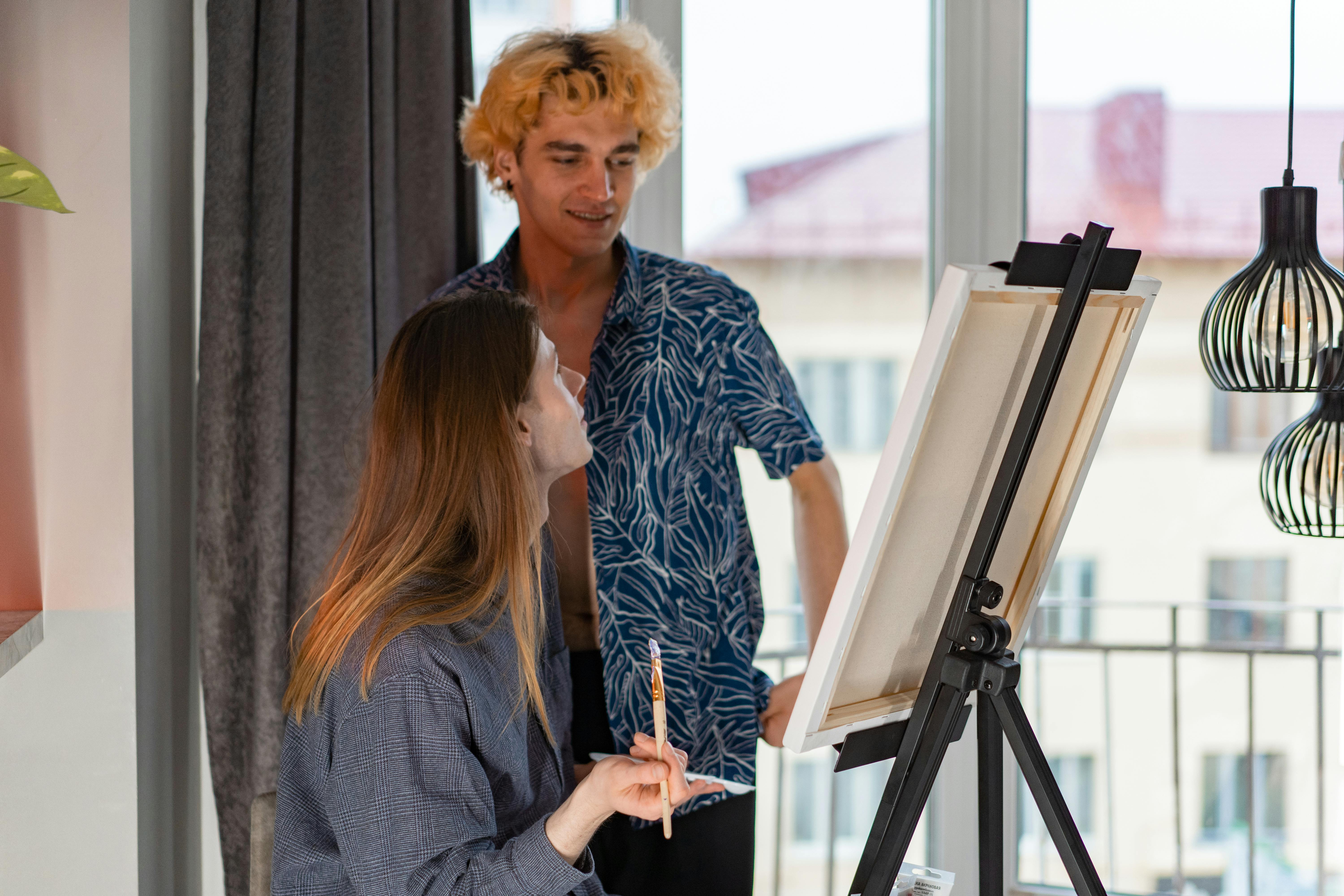 Two young adults engage in a lively painting session by large windows, creating artwork on an easel.