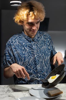 Young man cooking a fried egg at home for breakfast, enjoying a bright morning indoors.