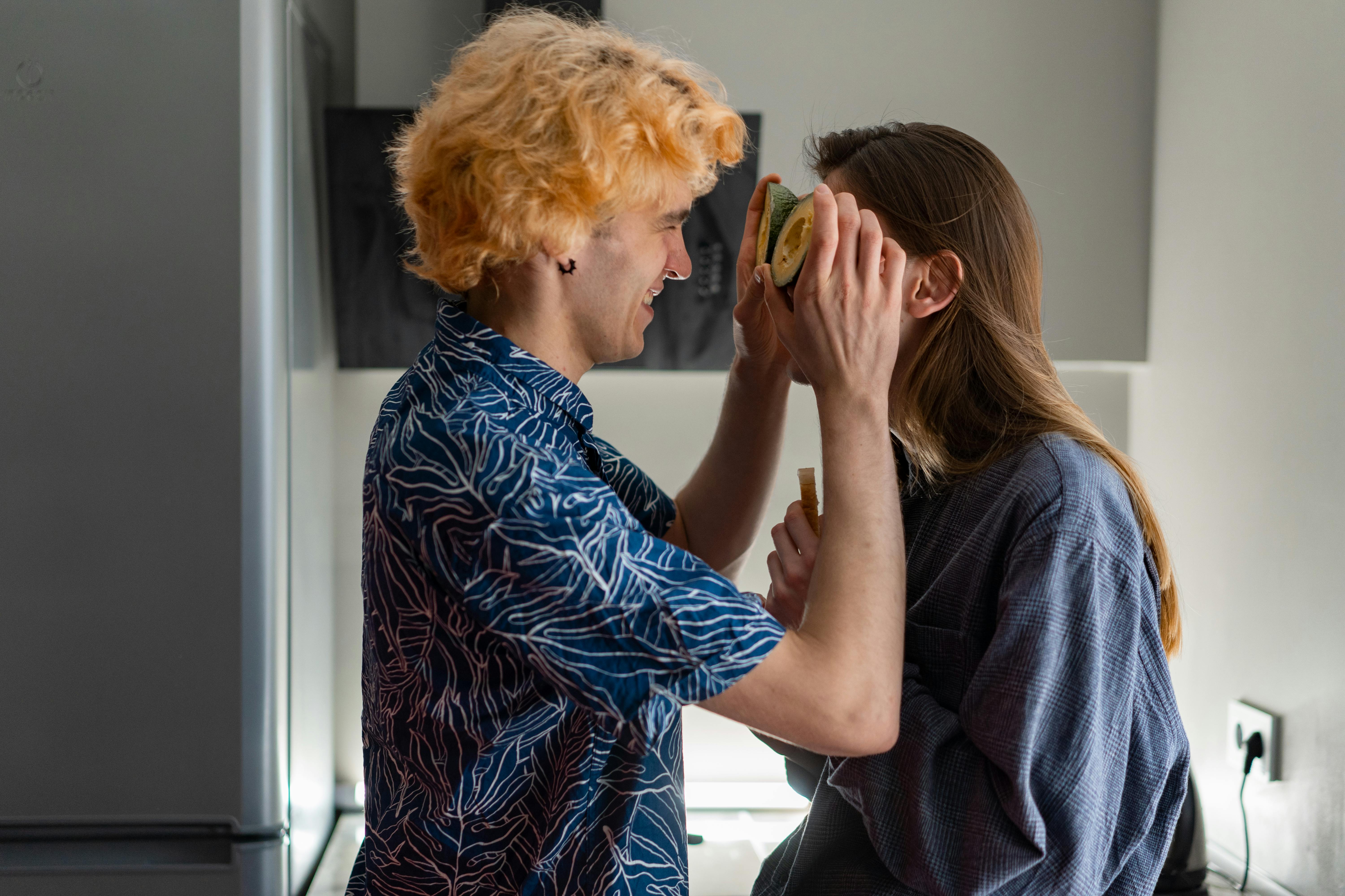 Playful couple in the kitchen, man holding avocado halves up to woman's face, both laughing at their silly moment together.